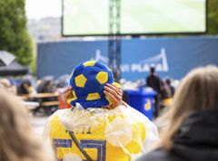 Person iklädd gul regnponcho och hatt med blå-gula fotbollsmönster tittar på en stor utomhusskärm.