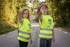 Två barn i reflexvästar står på en väg och gör stoppsignal.