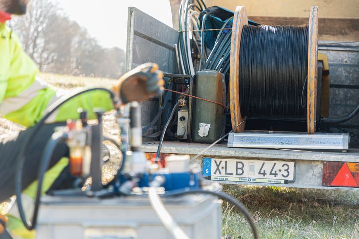 Foto som visar en lastbil, kablar och en hand i arbetet med att dra fiber. Landsbygd i bakgrunden