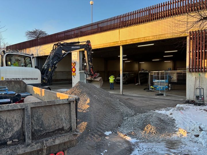 En grävskopa arbetar på byggarbetsplatsen medan en person i varselkläder står bredvid.