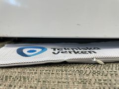 Närbild av en vit droppmatta med texten ”Tekniska Verken” som sticker ut under en diskmaskin på ett vävt köksgolv.