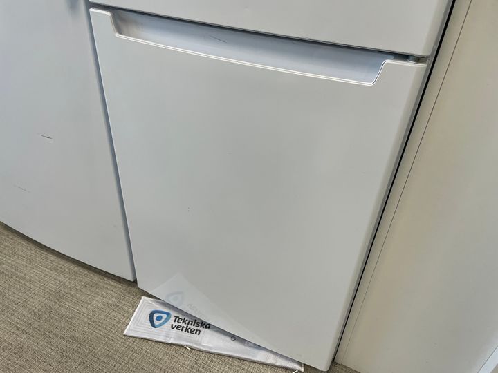 Ett vitt kylskåp i ett kök med en droppmatta som sticker ut undertill, märkt med logotypen “Tekniska Verken”.