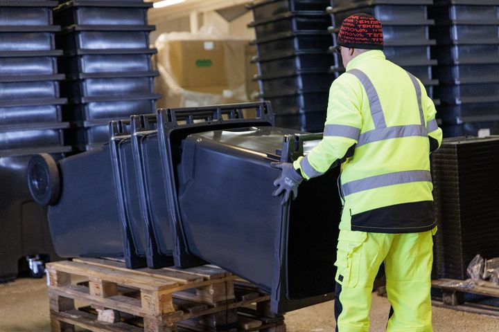 En person i varselkläder staplar svarta soptunnor på en pall i en industrilokal där soptunnor monteras.