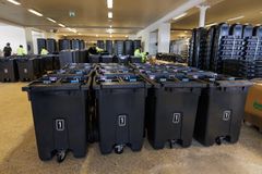 Svarta avfallskärl står uppställda i rader inomhus i en industrilokal medan personal i varselkläder arbetar med montering i bakgrunden.