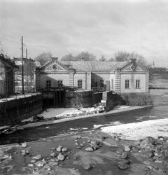 Svartvit bild av det gamla vattenverket i Tannefors vid ett delvis istäckt vattendrag. Den tegelklädda byggnaden står på en stenmur intill vattnet, med snö och is längs strandkanten.
