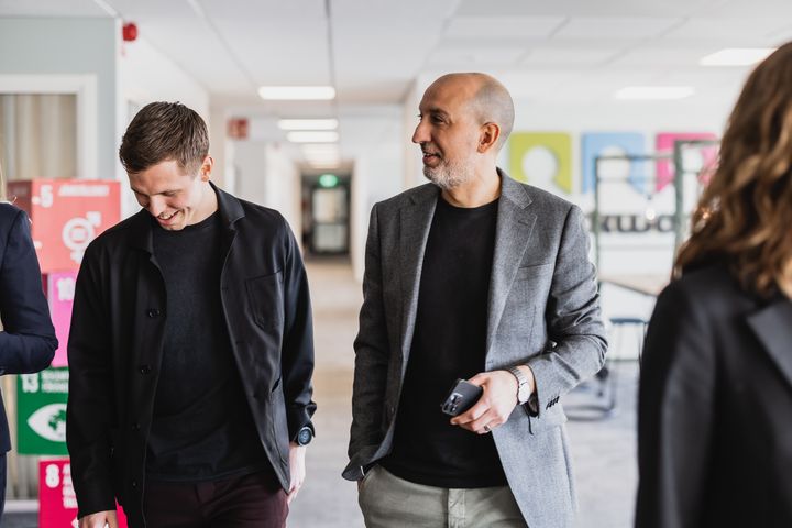 Två personer i affärsmiljö går tillsammans i en ljus kontorskorridor, klädda i kavaj och mörka kläder, med fokus på dialog och samarbete.