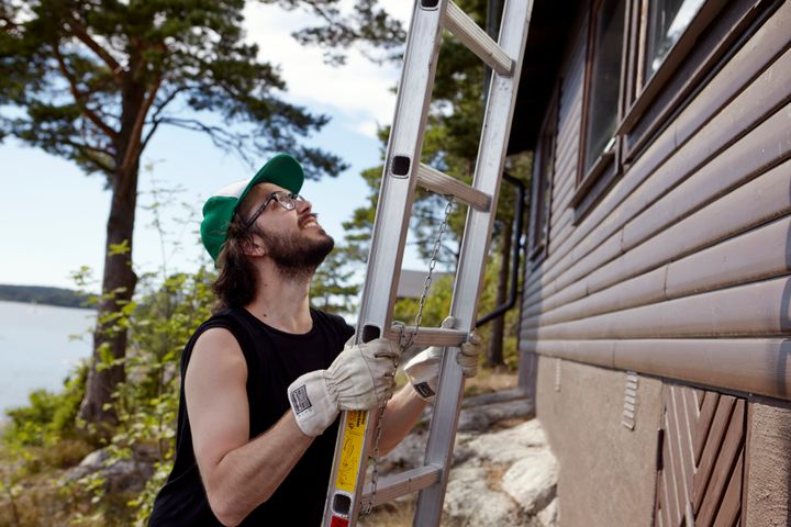 En man med glasögon och keps klättrar uppför en stege vid en träbyggnad utomhus.