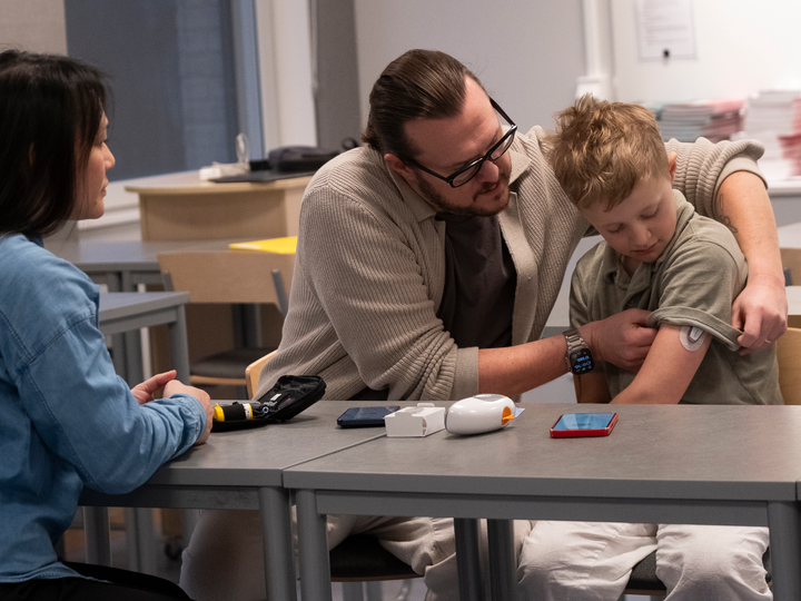Tre personer sitter vid ett bord i ett klassrum. En vuxen visar en annan vuxen hur man använder en medicinsk apparat på barnets arm.