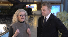 Två personer står och pratar inomhus, omgivna av moderna möbler.