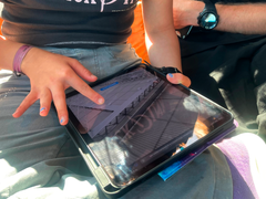 En person sitter med en Ipad i knät och klickar på något på skärmen. En annan person sitter jämte. Man ser bara händer, ben och armar.