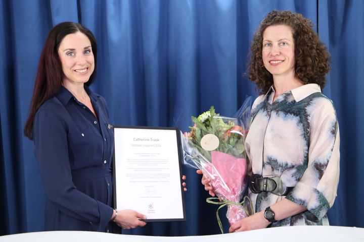 Två kvinnor står bredvid varandra, en tar emot ett diplom och en bukett blommor, leende mot kameran.