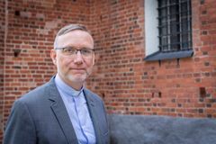 Man i kort hår, klädd i ljusblå prästskjorta utanför kyrkvägg av rött tegel