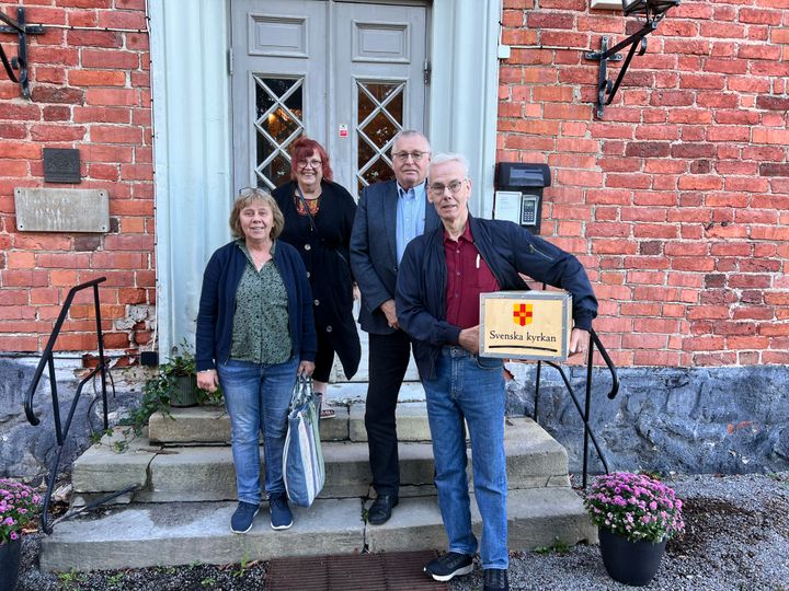 Fyra personer står framför en byggnad, en av dem håller en låda märkt "Svenska kyrkan".