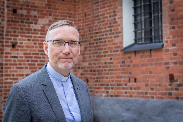 Man i kort hår, klädd i ljusblå prästskjorta utanför kyrkvägg av rött tegel