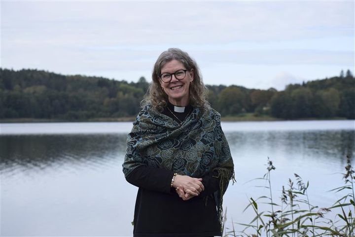 En kvinna i prästskjorta står leende framför en sjö med skog i bakgrunden.