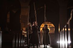 Tre personer i mörka kläder går genom en kyrka med höga stavar, scenen är dramatiskt belyst bakifrån.