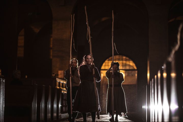 Tre personer i mörka kläder går genom en kyrka med höga stavar, scenen är dramatiskt belyst bakifrån.