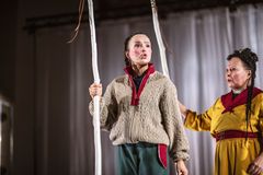 Två personer i traditionella dräkter står på en scen med vita trästavar.