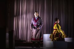 Två personer i färgglada dräkter uppträder på en scen med en mörk bakgrund. En person står medan den andra sitter.