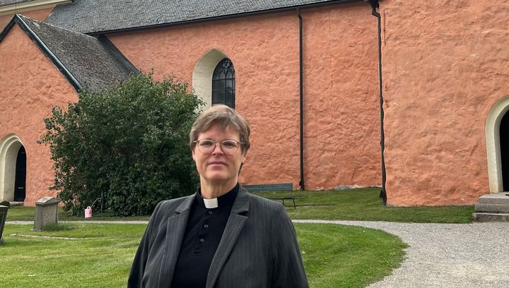 kyrkoherde Marjut Ervasti, kvinnlig präst framför en gammal kyrka med rosa väggar.