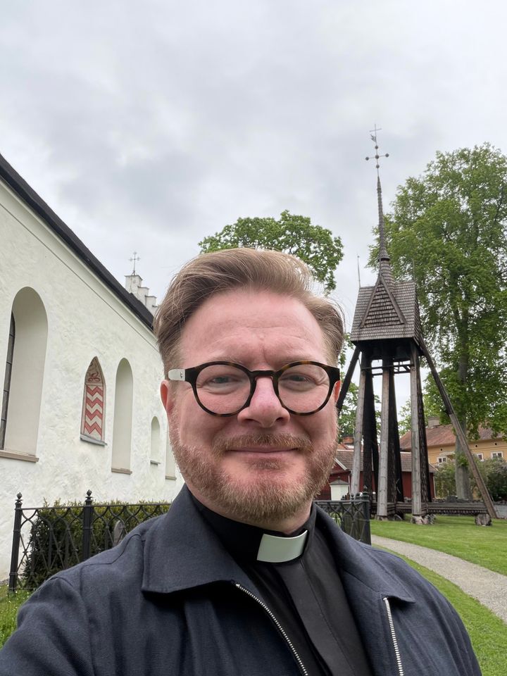 En man i prästkläder står framför en kyrkobyggnad och ett klocktorn.