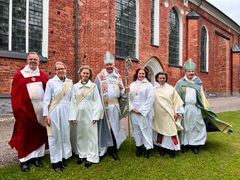 Sju personer: präster, diakoner och biskopar i liturgiska kläder framför katedral i rött tegel.