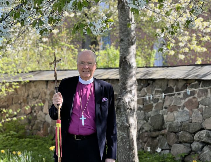 En man klädd i biskopsskjorta står utomhus, hållandes en vandringsstav. I bakgrunden är en stenmur och grönskande träd.
