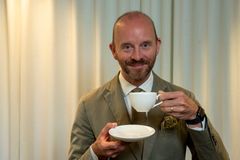 Porträtt av en leende man i kavaj som håller en vit kaffekopp och tefat, framför en ljus gardinbakgrund.