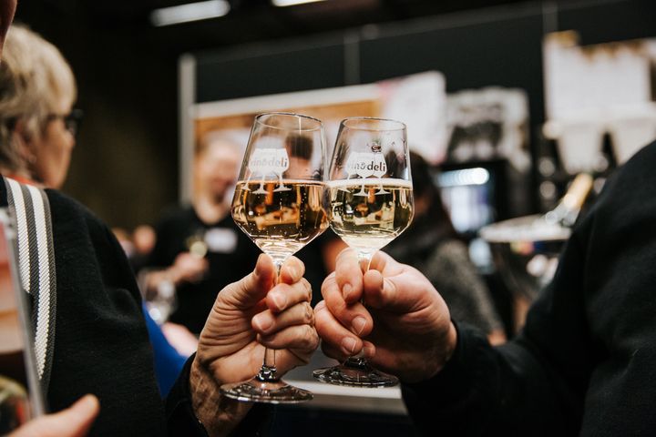 Två vinglas med vitt vin skålar mot varandra, hållna av besökare på en vinmässa i suddig bakgrund.
