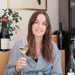 Leende kvinna med långt brunt hår håller ett glas vitt vin, omgiven av vinflaskor och en grön växt i bakgrunden.