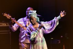 Angélique Kidjo sjunger på scen i mönstrad klänning, med armarna utsträckta och mikrofon i handen. En basgitarrist står bakom henne i bakgrunden.