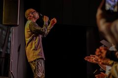 Youssou N’Dour i glasögon och skjorta sjunger i mikrofon på en scen, medan publiken till höger applåderar.