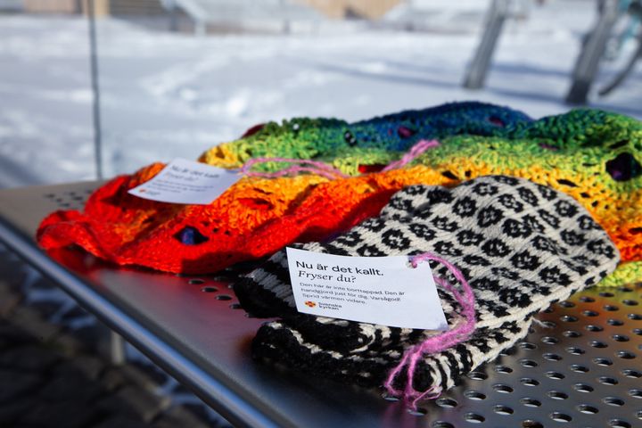 En färglad halsduk och svartvita vantar ligger i ett vindskydd. Den som behöver den kan använda plaggen för att värma sig.