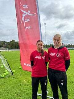 Två unga kvinnor i röda tröjor står bredvid varandra framför en flagga på en fotbollsplan.