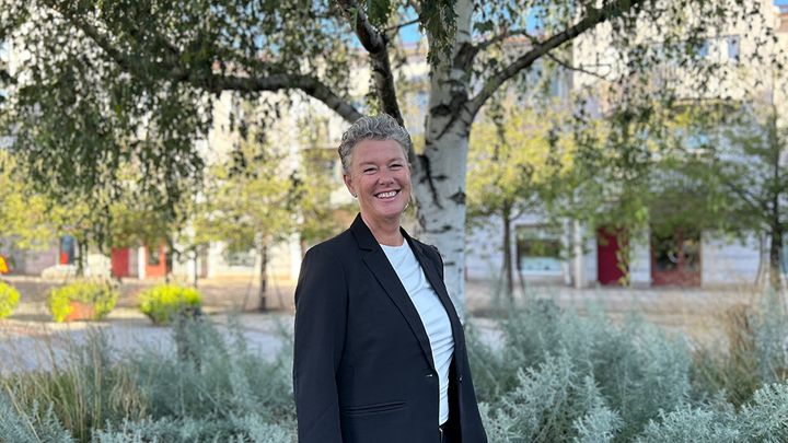 En person med kortklippt, ljust hår, vit t-shirt och svart kavaj står framför en björk och ler mot kameran. Ute lyser solen, björken är full av gröna blad och personen står i skuggan.