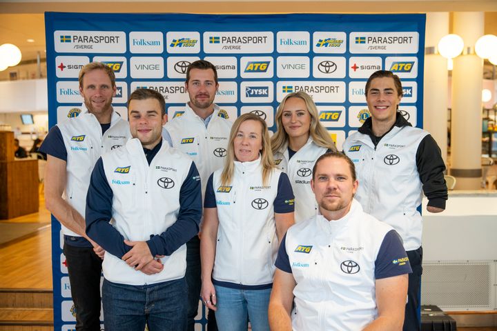 Gruppbild på de svenska paralandslagen i alpint och längd/skidskytte. Gruppen står framför en sponsorvägg.