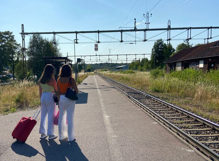 Två personer går på en perrong med resväskor i solskenet.