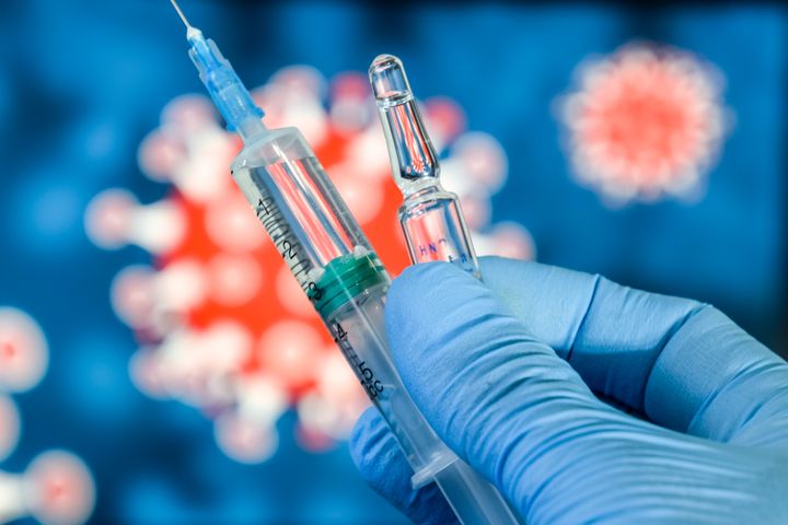 En spruta och en ampull hålls av en hand i en blå handske, med en suddig bild av ett virus i bakgrunden.
