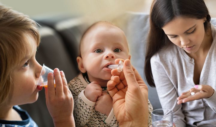 Barn som tar medicin; en vuxen hjälper en bebis och ett annat barn dricker från ett glas.