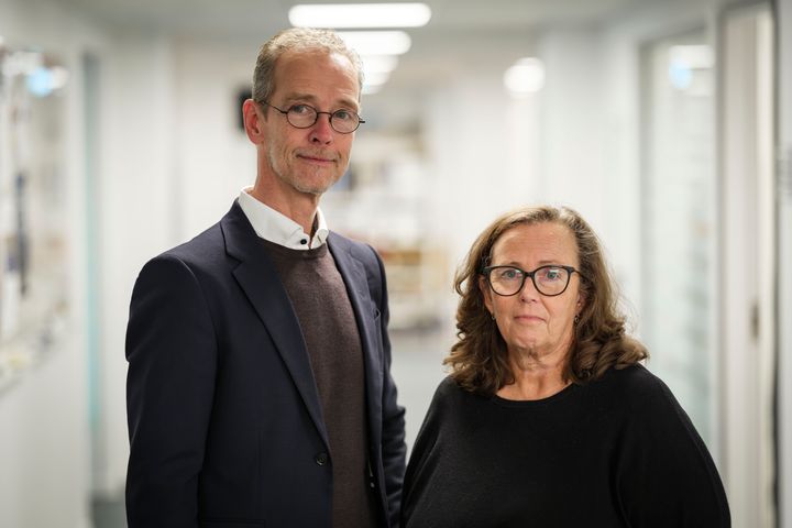 Två personer, Peter Asplund till vänster och Gunilla Andrew Nielsen, till höger står i en korridor. Den ena är klädd i en kostym och den andra i en svart tröja.