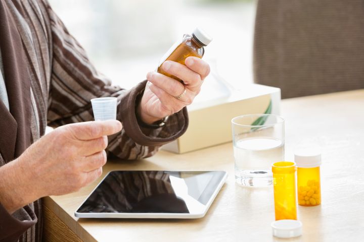 Person som håller en medicinflaska och en doskopp vid ett bord med en surfplatta, ett glas vatten och fler medicinflaskor.