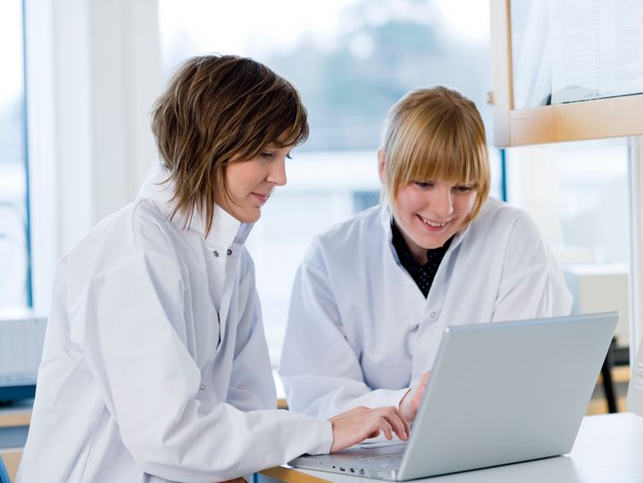 Två personer i vita labbrockar sitter vid en bärbar dator och arbetar tillsammans.