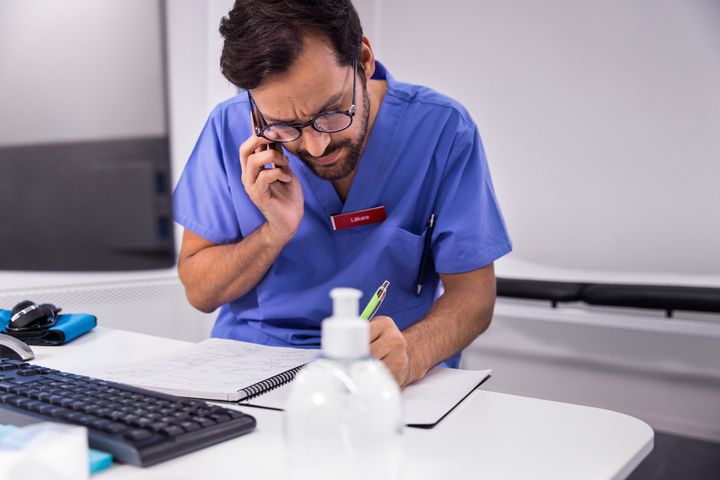 En vårdpersonal i blå uniform pratar i telefon och skriver anteckningar vid ett skrivbord.
