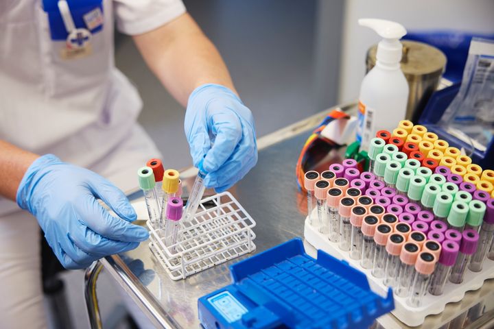 En vårdpersonal hanterar provrör med färgade lock på ett metallbord.