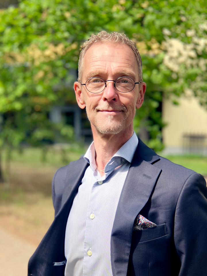 Peter Asplund i kostym står utomhus med träd i bakgrunden.