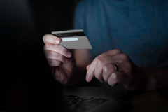 Person som håller ett kreditkort framför en dator, i ett svagt upplyst rum.