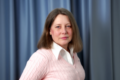 Bilden visar en kvinna i rosa tröja och vit skjorta framför en blå bakgrund.