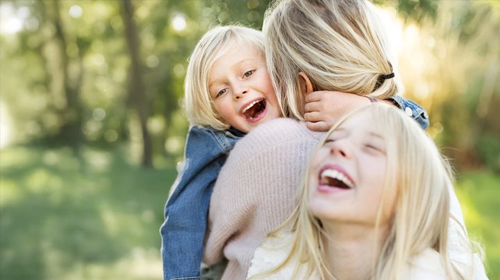 En kvinna och två barn skrattar och kramar varandra utomhus.