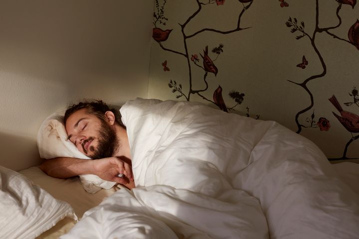 En person med skägg sover lugnt i en säng med vita lakan. En vägg med målade blommor och fåglar syns i bakgrunden.