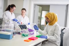 Tre personer arbetar i ett laboratorium med pipetter och färgade provplattor. En person bär en gul sjal.